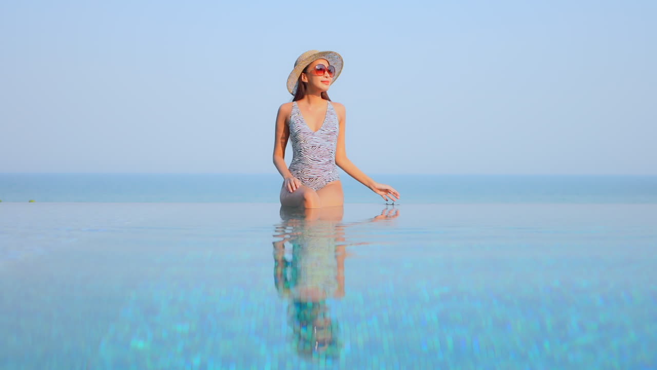 красивая женщина в купальнике сидит на бортике бесконечного бассейна, касаясь своей шляпы от солнца и подняв руки вверх с потрясающим видом на горизонт тропического моря в пхукете, таиланд, статическое пространство копий