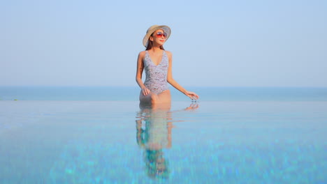 pretty woman in swimwear sitting on infinity pool border, touching her sunhat and raising hands up with a stunning view of tropical sea horizon in phuket, thailand, static copy space
