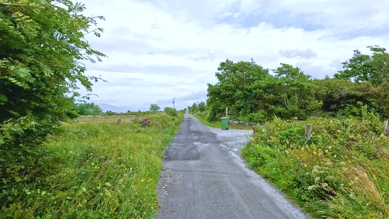A vehicle travels along a narrow, winding country road bordered by lush greenery and fields under overcast daylight, captured in smooth forward motion