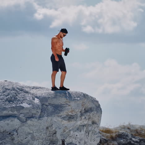 muscular male in shorts only drinking water after hard workout outdoors. bodybuilder standing on white rock with flask on the sky background.