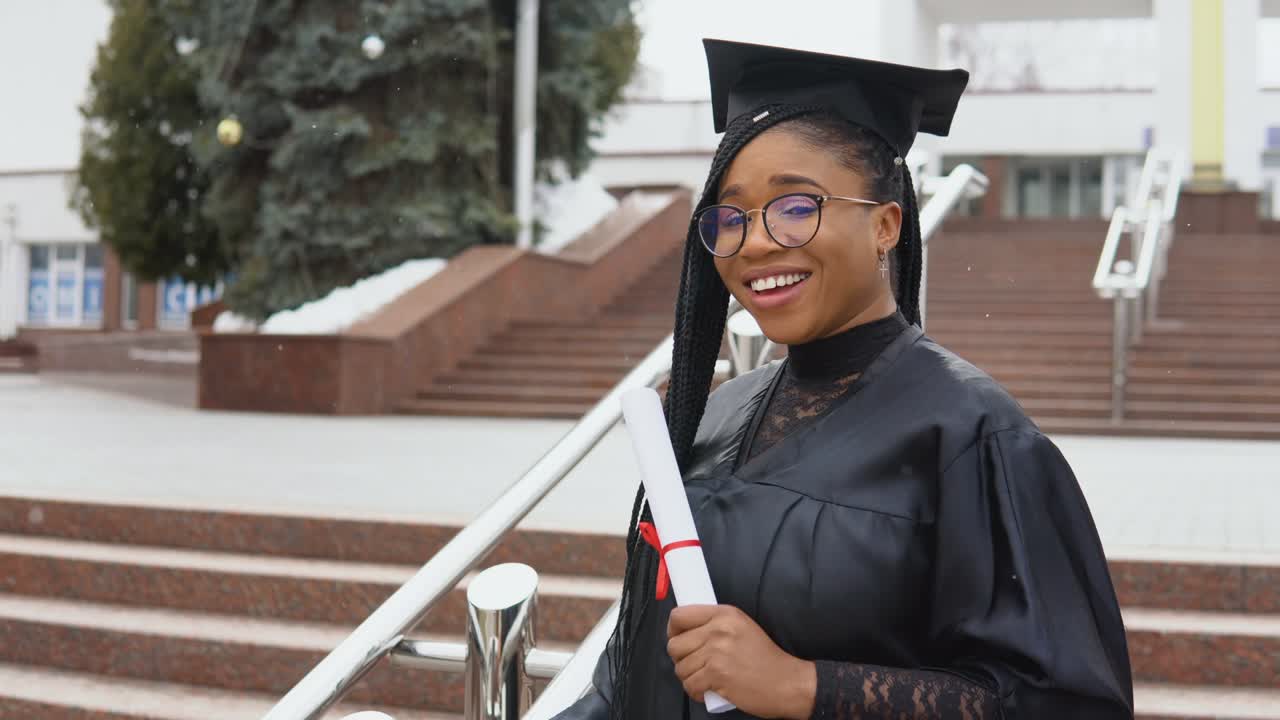 una mujer joven en la universidad en el manto del maestro sostiene un diploma y mira a la cámara. retrato con las escaleras frente a la entrada central de la universidad en fondo
