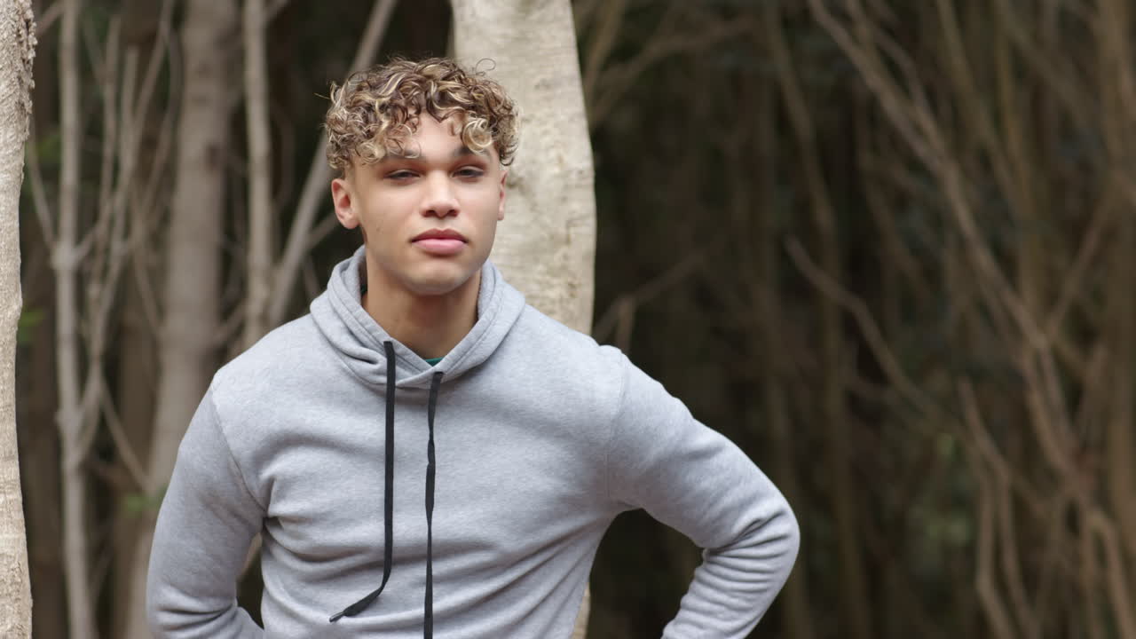 Young man in gray hoodie standing outdoors, looking thoughtful in forest