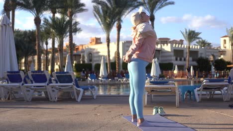 Young-adult-female-person-does-gymnastic-exercising.-Rise-up-bent-leg-on-bush-with-green-leaves-background.-Yoga-pose-near-poolside-in-sunny-day-time.-Healthy-lifestyle,-workout-therapy-outside.