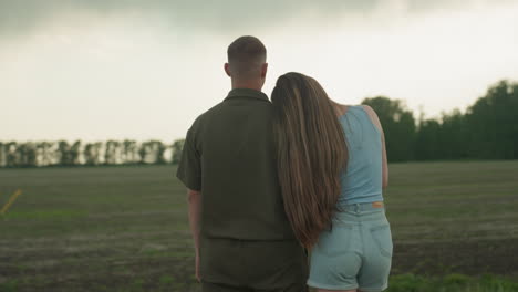 man and woman stand on serene rural farmland edge while man gestures toward horizon and woman rests head on his shoulder under dramatic cloudy sky with soft light