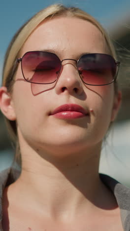 mujer blanca con camisa gris y gafas de sol lleva la bolsa de compras hacia atrás con una expresión enfocada, está de pie al aire libre con un fondo borroso, y se ve a dos personas pasando detrás de ella