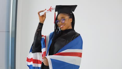 happy stylish afro american woman student in graduate uniform shows diploma and smiling with british flag on shoulders