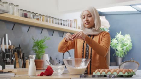 Video-De-Una-Mujer-Birracial-Feliz-Con-Hijab-Horneando-En-La-Cocina-De-Casa,-Rompiendo-El-Huevo-En-Un-Tazón