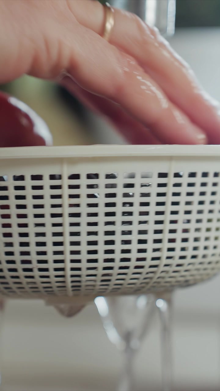 Close up of hand with gold ring washing white plastic colander. Hand moves colander and its contents water drips steadily. Colander is held over glasses as water drips focusing