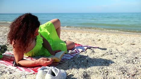 chica caucásica de pelo rojo con vestido verde se relaja al sol en la playa leyendo un libro con la brisa moviendo su pelo rojo, tiro estático