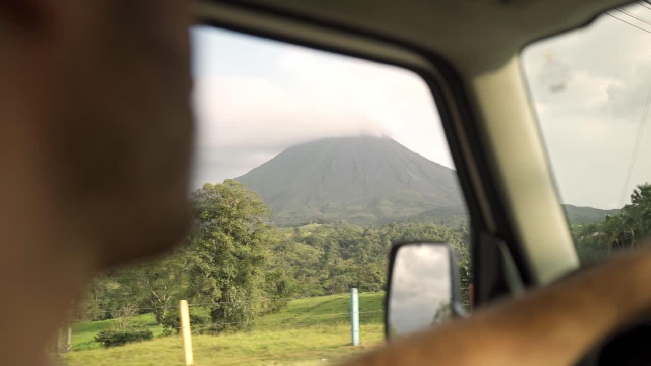 Interior view of Man driving a car with Arenal Volcano at sunset in the background in Costa Rica