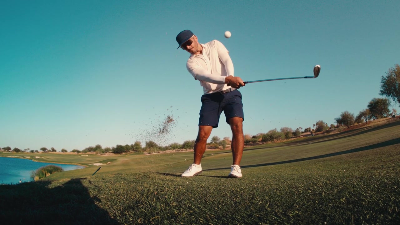A golfer flushes a wedge shot on tight fairway lie and watches the ball fly under a clear blue sky, captured in slow motion. Strong for premium golf and lifestyle content.