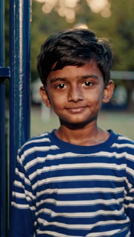 portrait of a confident young boy wearing a blue and white striped t shirt, standing near a blue metal fence, illuminated by the warm glow of golden hour