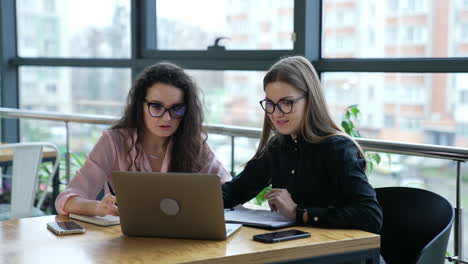two women colleagues discuss something in computer in office. female teammates wearing glasses work in front of laptop.