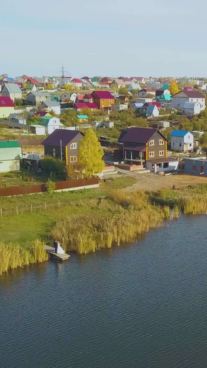 distant couple on small wooden pier near large river against neat cottage town on nice autumn day aerial view