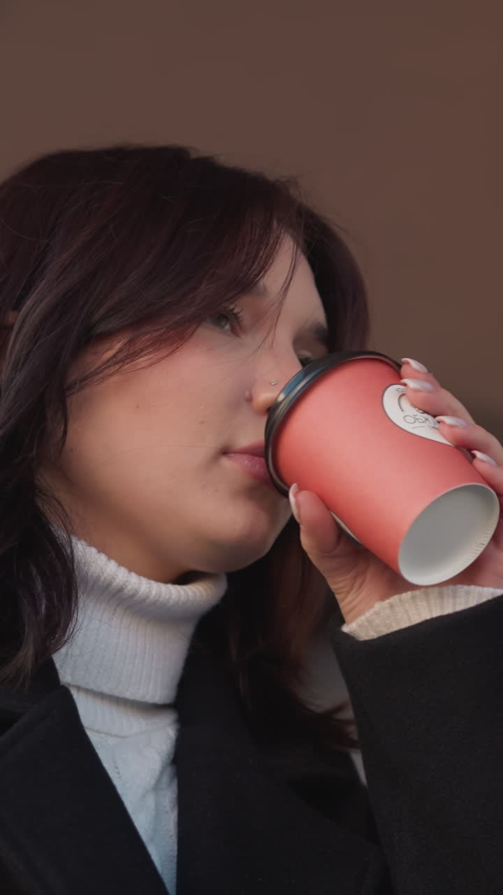 검은 코트를 입은 여자가 마당 에서 차 컵을 들어 올리고, 한 잔을 마시기 위해 멈추고, 웃음을 터뜨리고, 컵을 테이블 위에 부드럽게 다시 놓고, 앞에는 부분적인 노트북이 보인다.