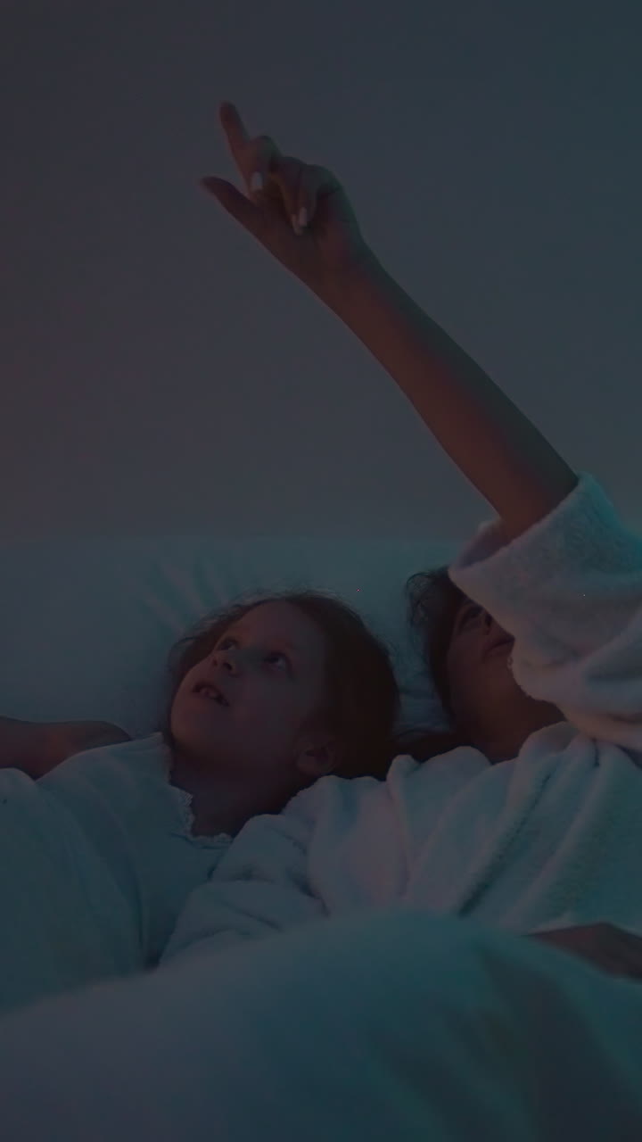 아이들과 함께 편안한 침대에 누워있는 젊은 여성은 벽에 다채로운 별을 반사하는 프로젝터를 가지고 있습니다. 배경에 별이 있는 밤 하늘을 가진 어린이를 위한 공간