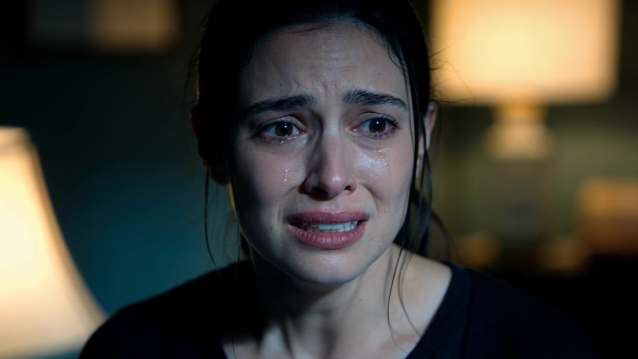 Una donna che piange con un top nero fissa la telecamera, versando lacrime, con il labbro tremante, reagendo al dolore, con lampade sullo sfondo.