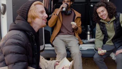 un equipo de producción de películas tomando un descanso mientras come un sándwich y habla detrás de una camioneta en la calle 3