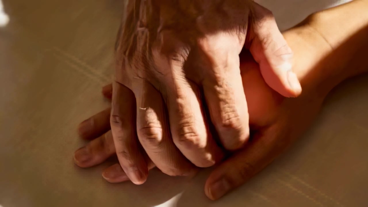 Shifting senior hand pressing and covering adult hand on pale sheet as sun shifts, giving comfort