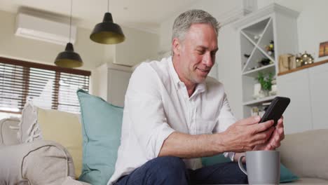 Happy-caucasian-man-sitting-on-sofa-in-living-room,-using-smartphone