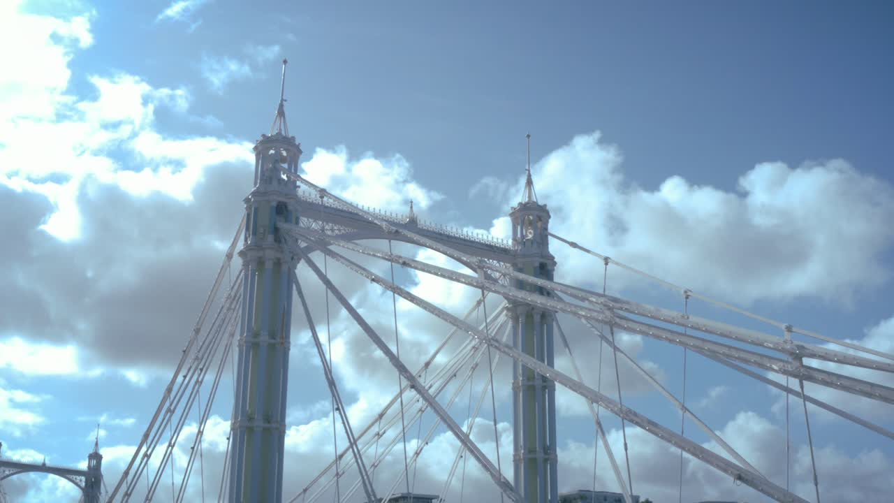 Close-up aerial view of the intricate structural details of Albert Bridge in Chelsea, London. The shot highlights the colourful suspension cables, steel framework, and decorative Victorian engineering