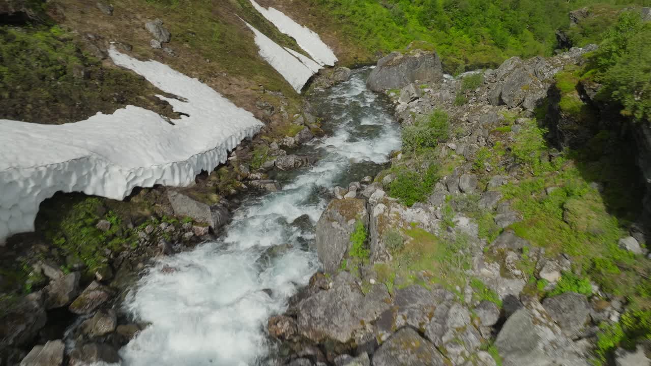 Droneoptagelser, der følger et vandløb omkranset af sne langs bredderne og frodigt grønt terræn, hvilket fanger en slående kontrast mellem vinter og forår i et naturskønt landskab
