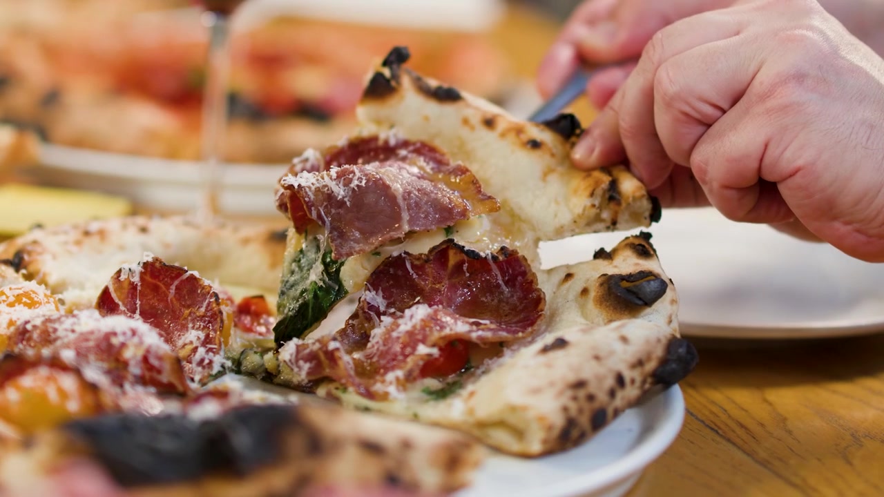 Primo piano di mani che tagliano una pizza al prosciutto con coltello e forchetta durante la cena.