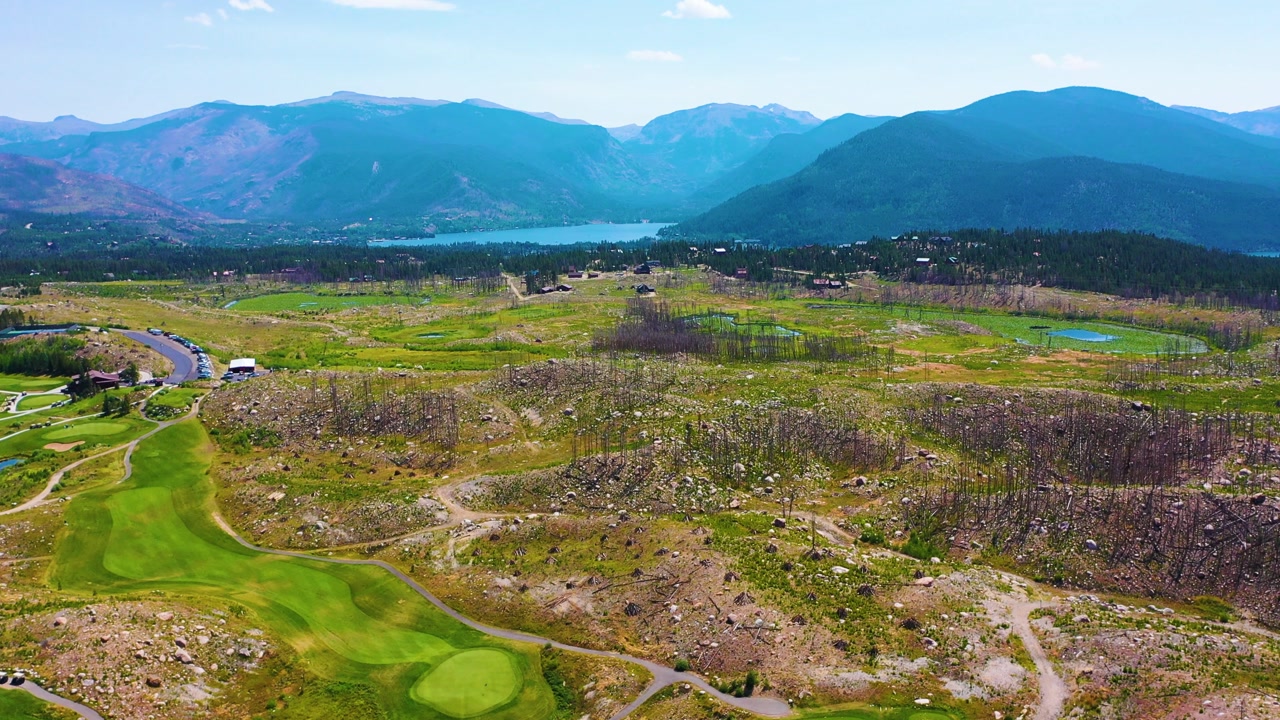 Expansive drone view of mountain golf fairways crossing post wildfire wilderness, with charred slopes, rocky hills, sparse vegetation, and dramatic ridgelines stretching across the background.