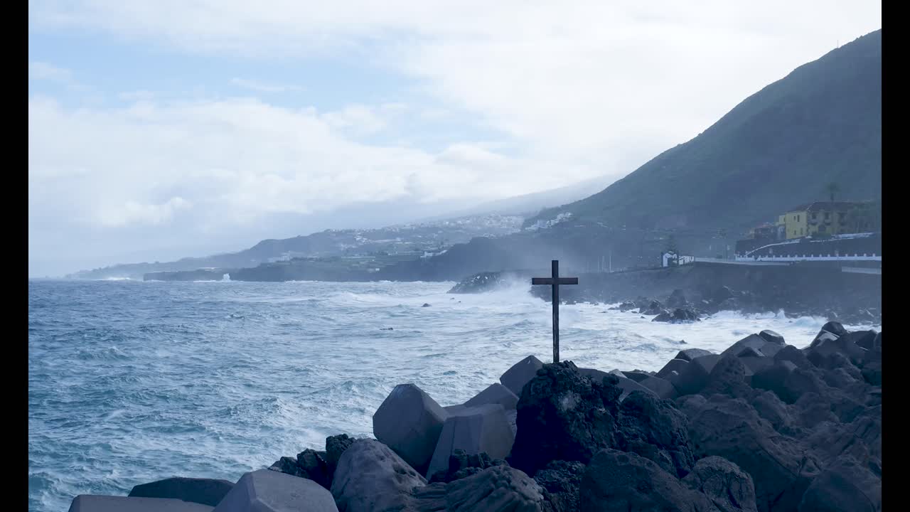 Croce su costa rocciosa con onde oceaniche impetuose e una città in lontananza