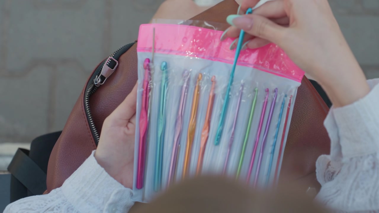 Primo piano di una giovane donna che tiene uncinetti colorati in confezione di plastica, poi li ripone in una borsa marrone con decorazioni esagonali. La scena è ambientata all'aperto su pavimentazione grigia sotto una luce soffusa.