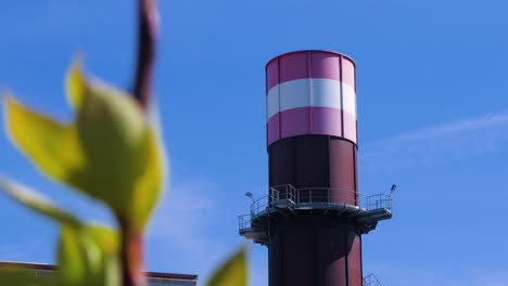 Exterior-view-of-abandoned-Soviet-heavy-metallurgy-melting-factory-Liepajas-Metalurgs-territory,-rust-covered-chimney,-out-of-focus-tree-in-foreground,-sunny-day,-medium-shot