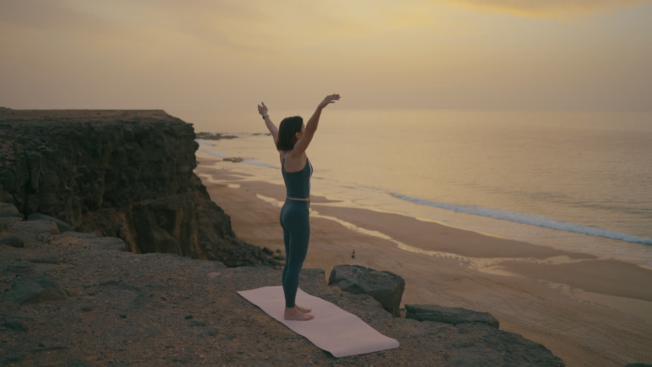 Woman practicing yoga on a cliff overlooking the ocean at sunset.