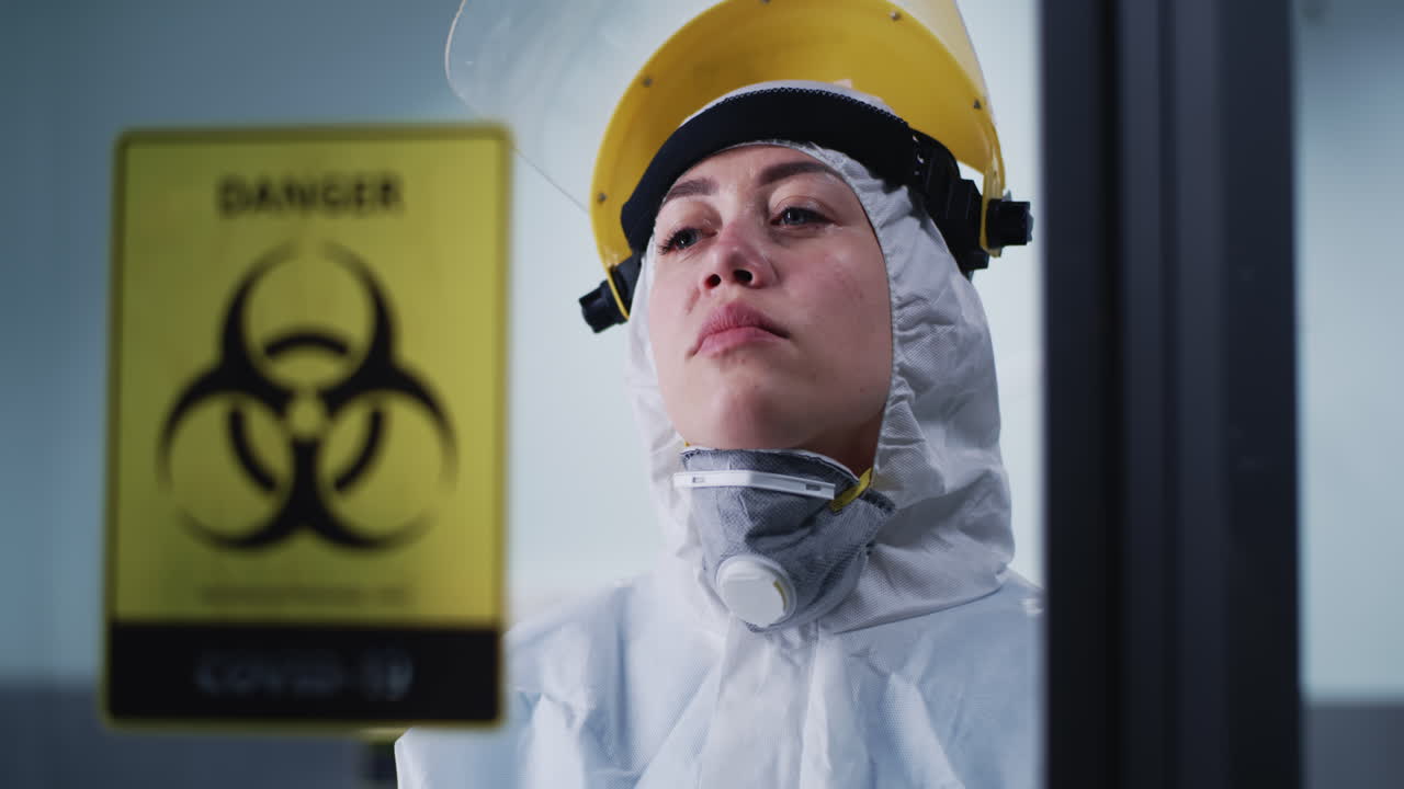 A healthcare worker or scientist in a hazmat suit and face shield with a distressed expression, next to a 'Danger Biohazard COVID-19' sign