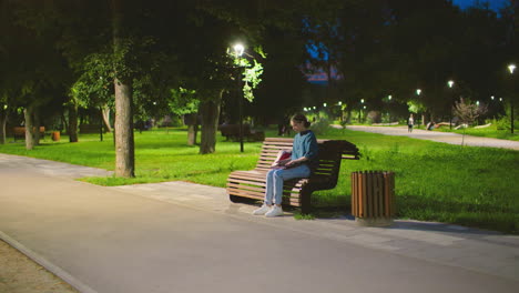 young woman sitting on bench outdoors opens laptop in peaceful park setting at dawn, light poles illuminate green landscape with people strolling in background