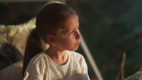 sunlit child looks at sunrise in room. thoughtful little girl looks at nature outside window. pretty kid rests sitting in soft chair on terrace