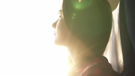 low angle shooting. the young woman is going by train. the face is flooded with sun rays. looks out the window, puts on the earphones. long haired caucasian