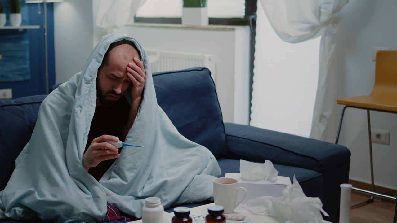 Ill person wrapped in blanket looking at thermometer