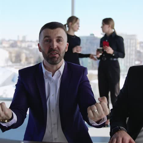 executive speaking emotionally on the camera and gesturing with his hands. two businessman sitting next to each other talking and smiling close up. two female office workers drinking coffee at the background