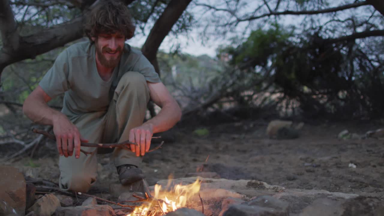 Happy bearded caucasian male survivalist adding kindling to fire at camp in wilderness