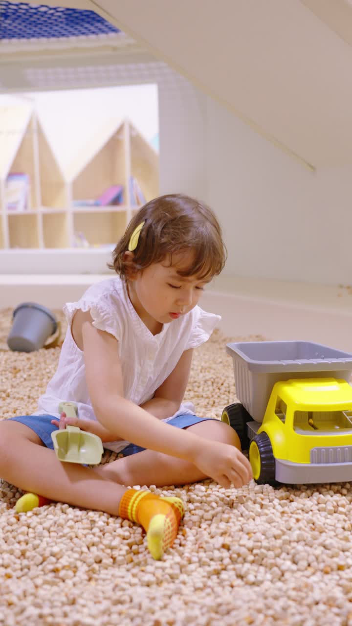 A cute little girl playing with a toy dump truck and bucket on an indoor playground filled with small granules