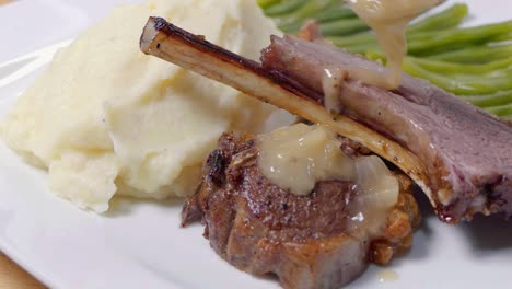 slider shot of adding sauce to lamb cutlets on a white plate with mashed potato and green beans