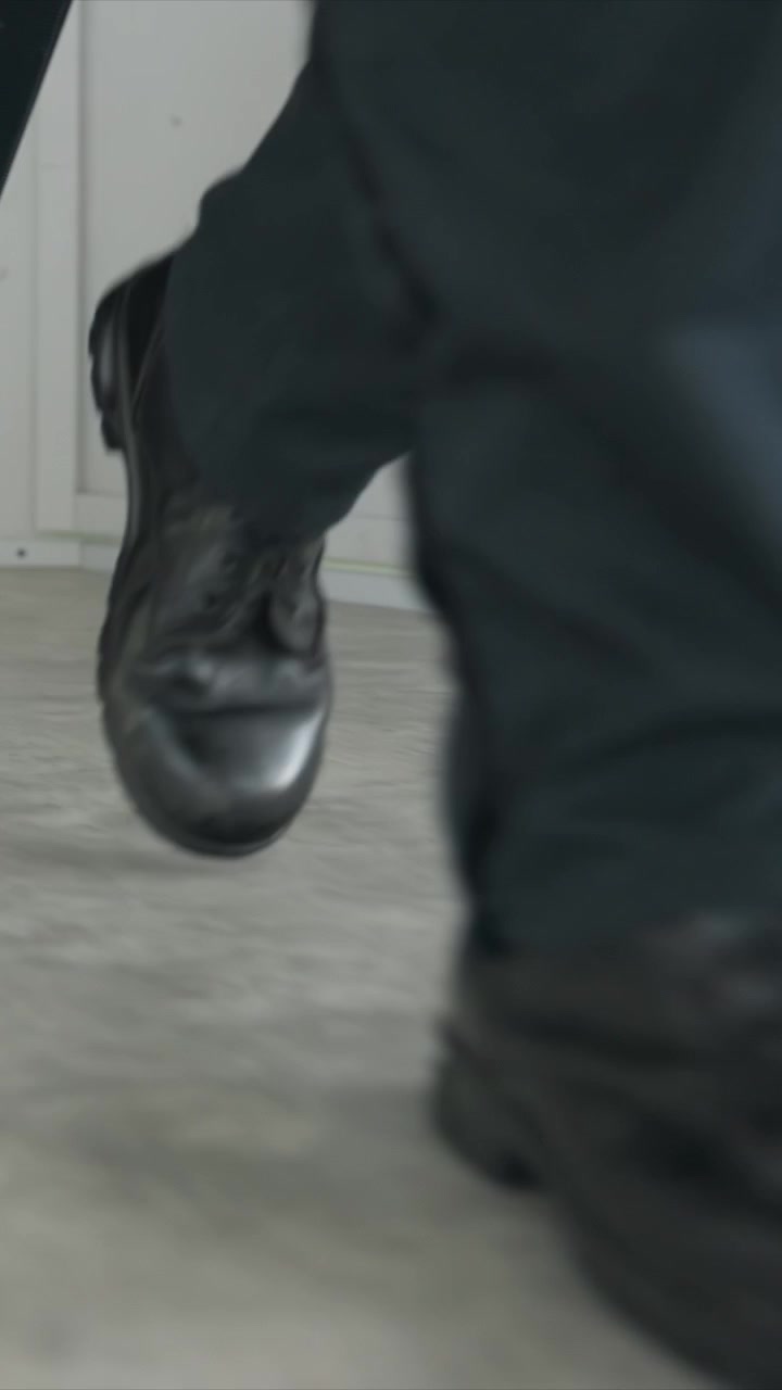 Primo piano dei piedi di un uomo con scarpe nere e pantaloni scuri che cammina. L'uomo continua a muoversi in avanti all'interno, la telecamera segue in basso. L'ambiente interno presenta pareti bianche e di colore chiaro.