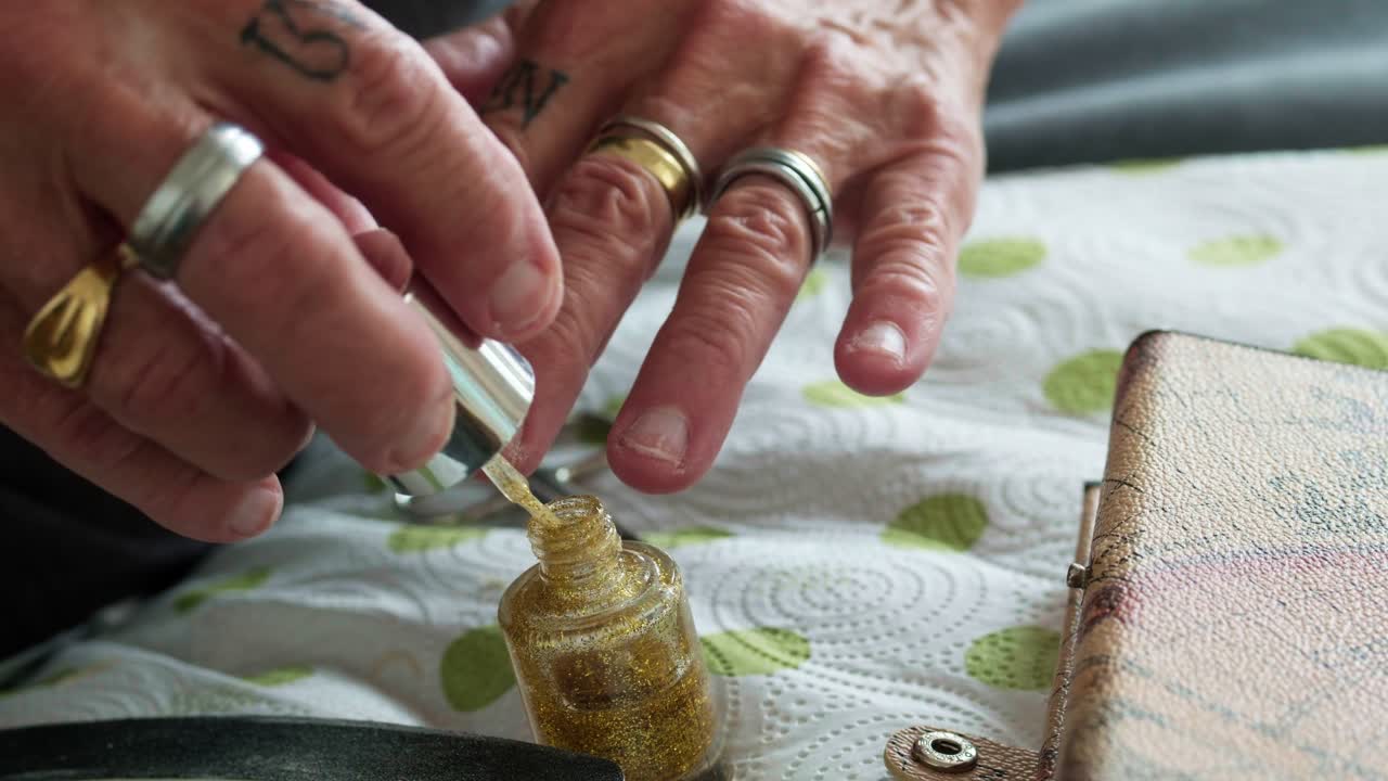 Close-up of senior hands applying gold glitter nail polish during a DIY home manicure. Detailed shot of beauty care routine featuring rings, nail tools, and shimmering lacquer for a festive look