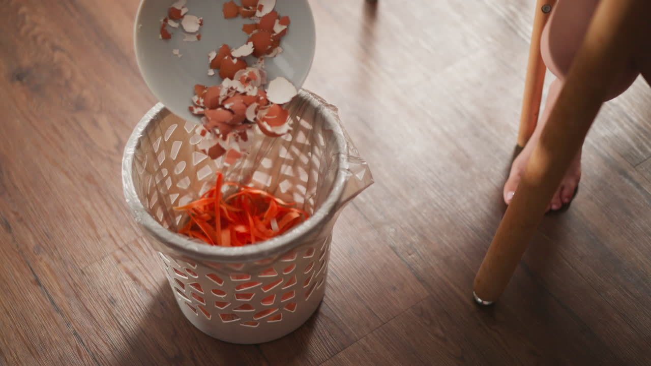 waste bin filled with vibrant carrot peels lined with plastic bag placed on polished wooden floor with partial view of seated barefoot person disposing egg shells into bin