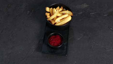 french fries with salt next to a mug of ketchup are presented on a rotating plate