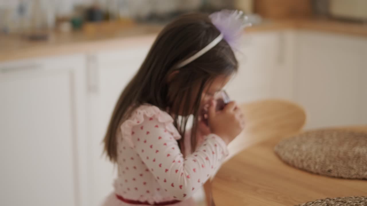 una niña toma un sorbo de un vaso mientras está sentada en una mesa de madera. lleva una camiseta de mangas largas con un patrón de corazón y una cinta para la cabeza, disfrutando de un momento tranquilo en un ambiente cálido y acogedor del hogar.
