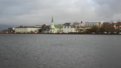 Hallgrimskirkja-Church-with-Green-Spire-Sits-on-Waters-Edge-of-Tjornin-Pond-Surrounded-by-Reykjavik-City-on-Overcast-Day,-Drone-Aerial