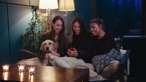 Three-girls-are-sitting-on-the-sofa-and-talking-in-a-cozy-evening-room,-next-to-them-is-their-light-colored-dog.-Candles-on-the-table-and-yellow-lamp-lighting