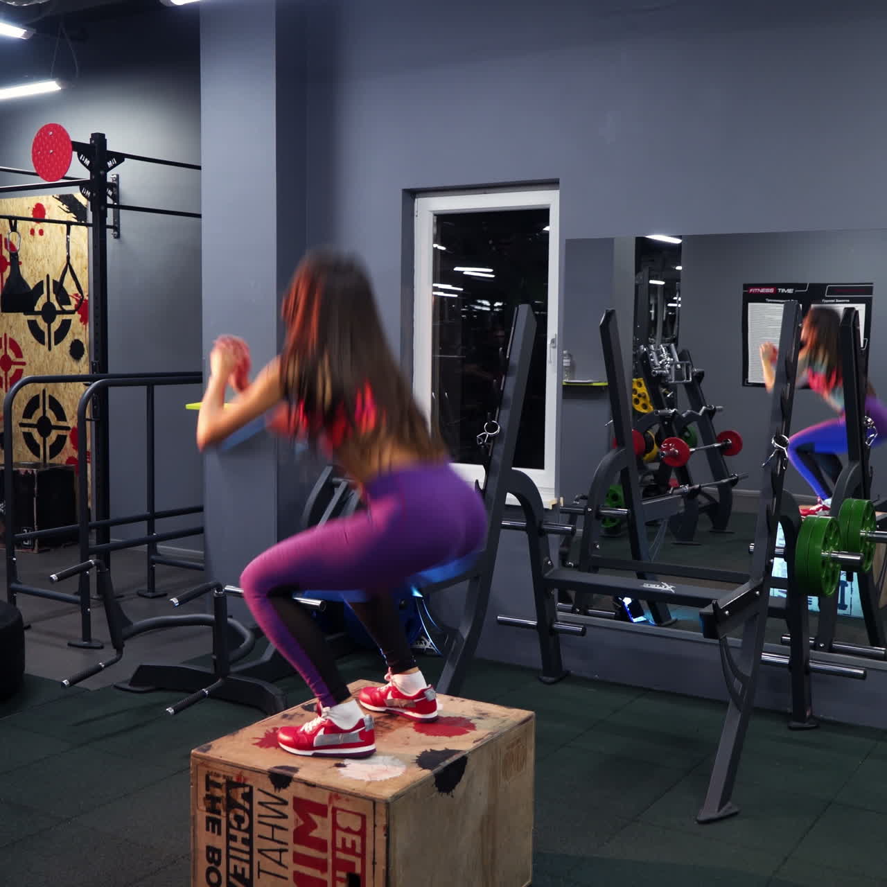 Athletic woman in purple sportswear does jumps on a big box in modern fitness gym. Intense exercise jumping of a young female. Side view of a woman jumping during her daily workout.