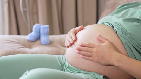 Caring future mother stroking her big tummy while resting on the sofa. Woman caressing her child in the big belly. Two little baby socks are on the armrest at he backdrop.